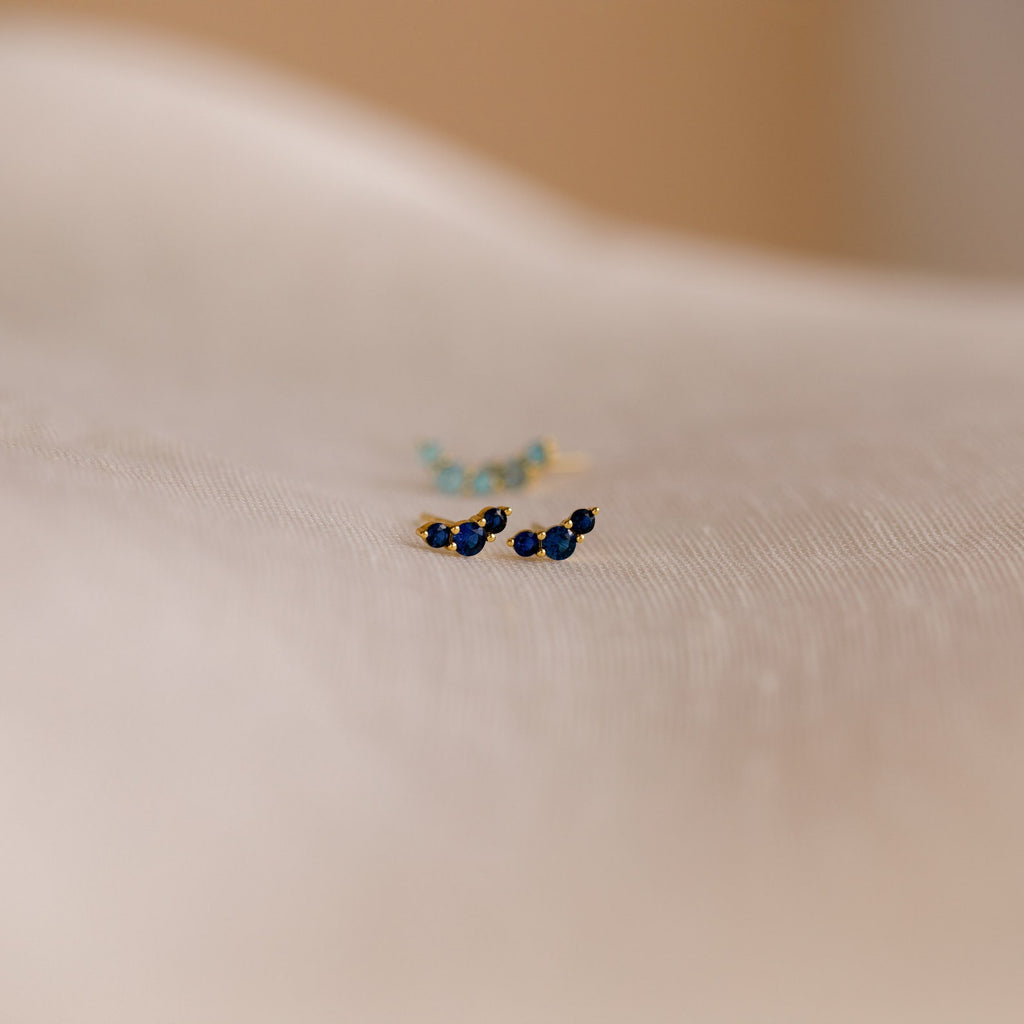 Bella Birthstone Studs, featuring small gold settings with blue gemstones, rest on a soft, light-colored fabric—an elegant gift for mothers and daughters.