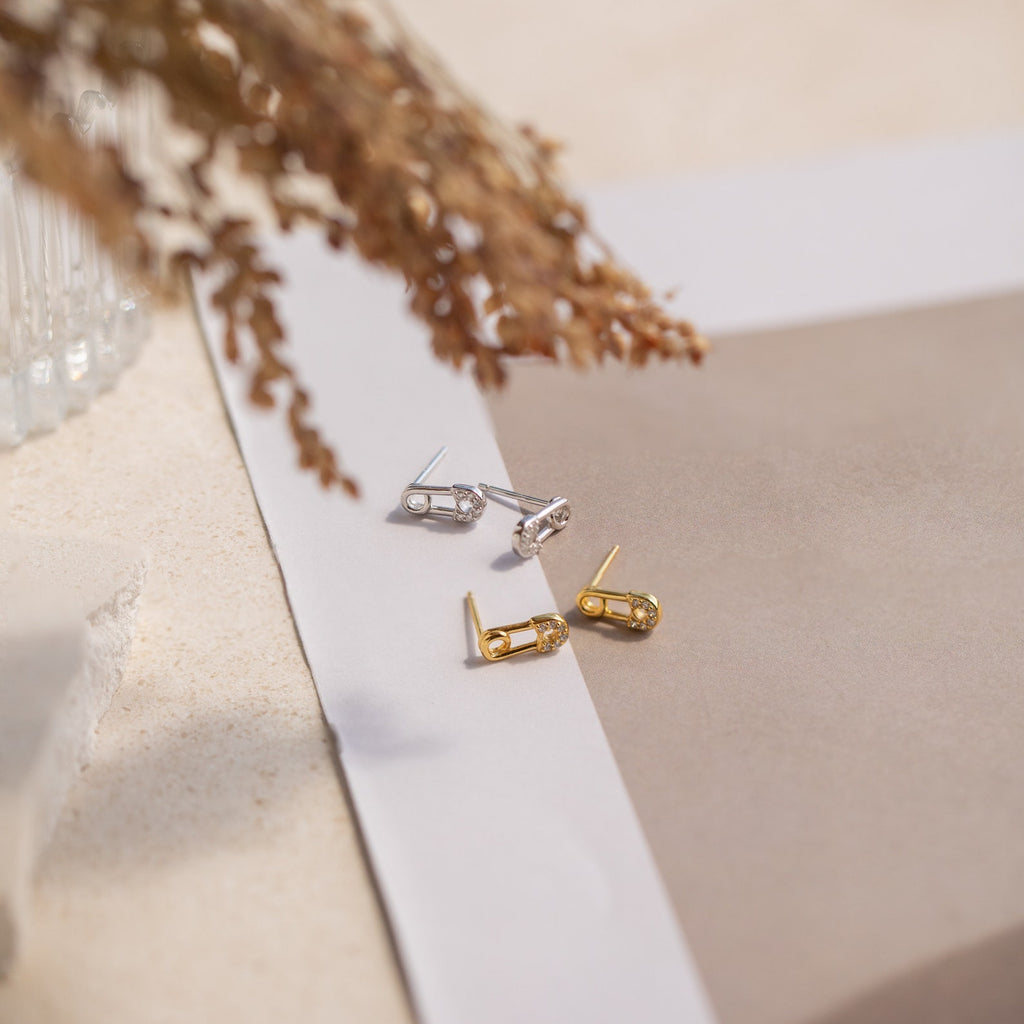 Two pairs of stud earrings, including the Tiny Pave Safety Pin Studs in silver and a gold CZ diamond stud, are displayed on neutral paper with dried foliage nearby.