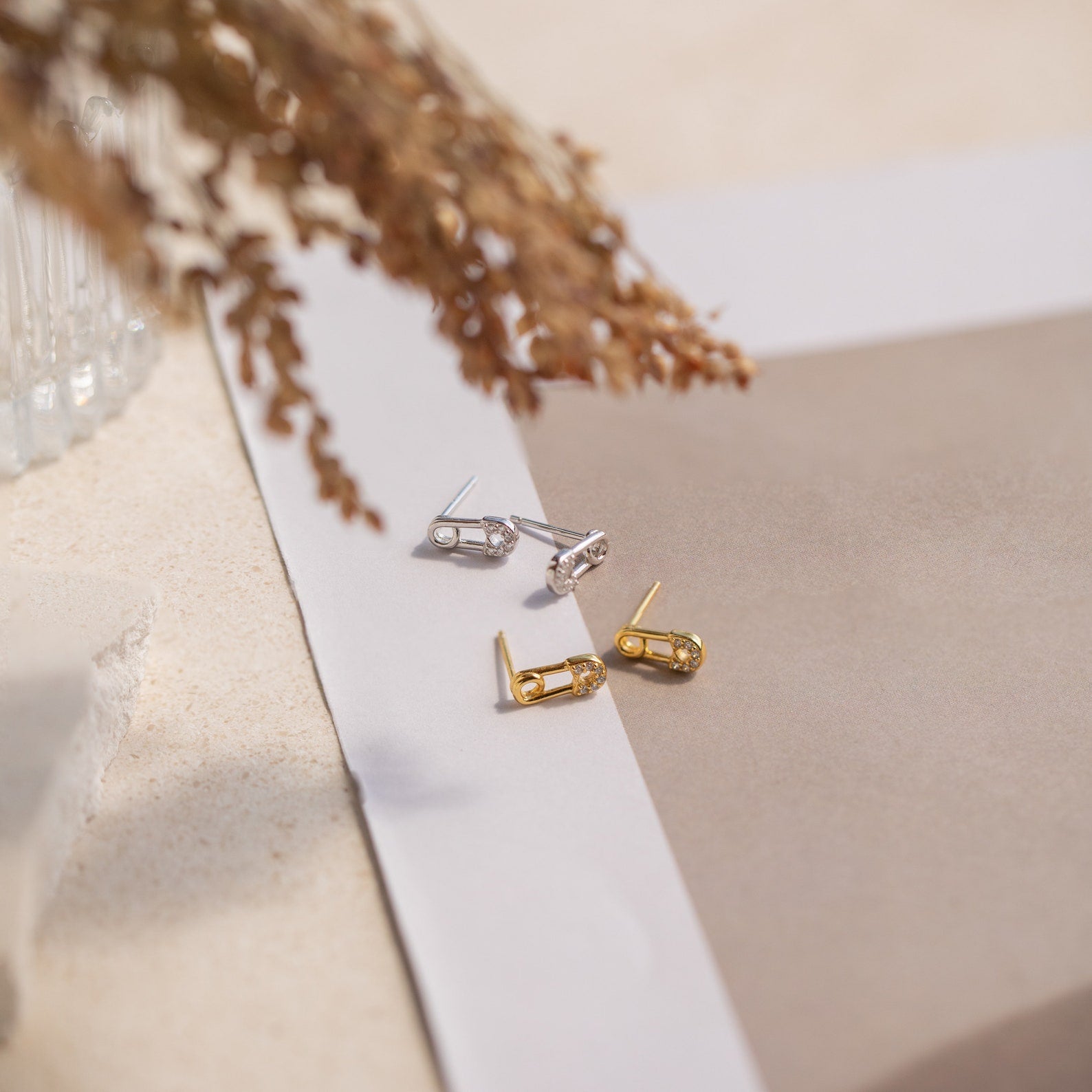 Two pairs of stud earrings, including the Tiny Pave Safety Pin Studs in silver and a gold CZ diamond stud, are displayed on neutral paper with dried foliage nearby.