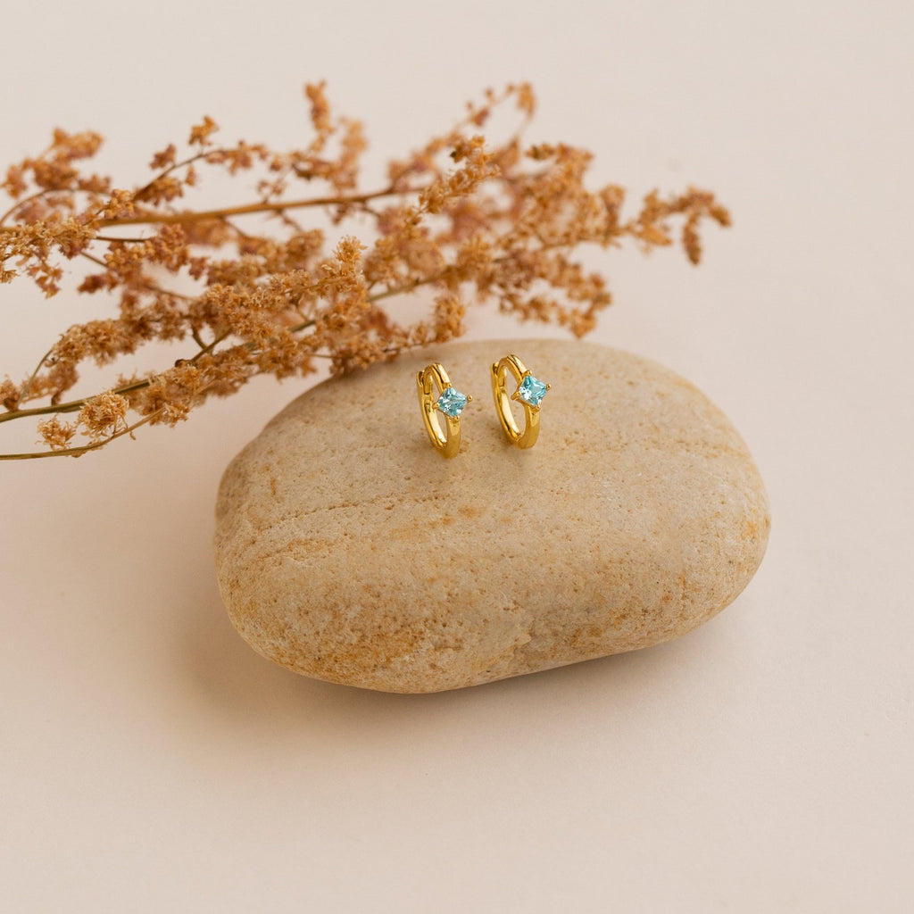Athena Opal Huggies—gold hoop earrings with blue opal gemstones—displayed on a smooth stone, accented by dried flowers in the background.