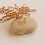Athena Opal Huggies—gold hoop earrings with blue opal gemstones—displayed on a smooth stone, accented by dried flowers in the background.