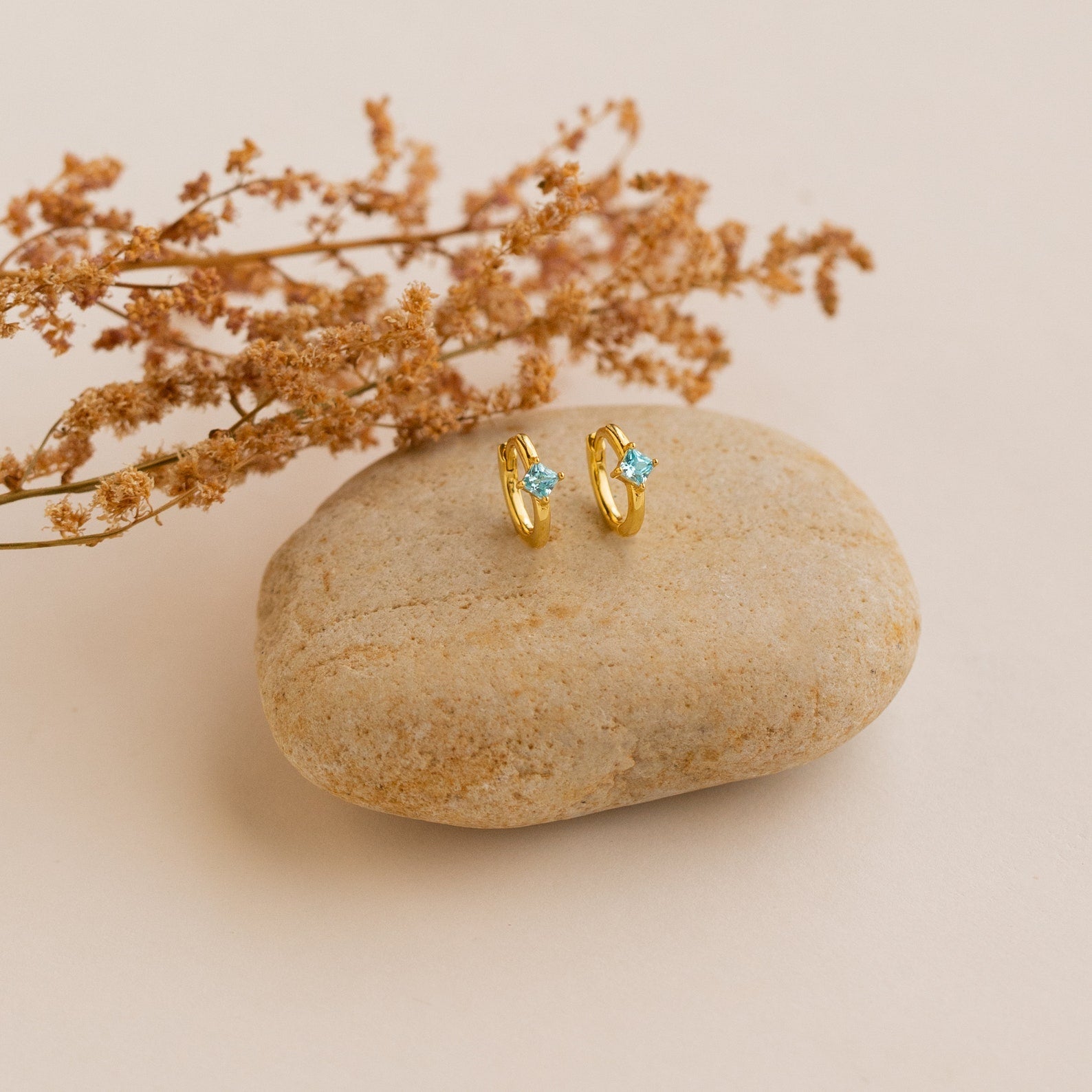 Athena Opal Huggies—gold hoop earrings with blue opal gemstones—displayed on a smooth stone, accented by dried flowers in the background.
