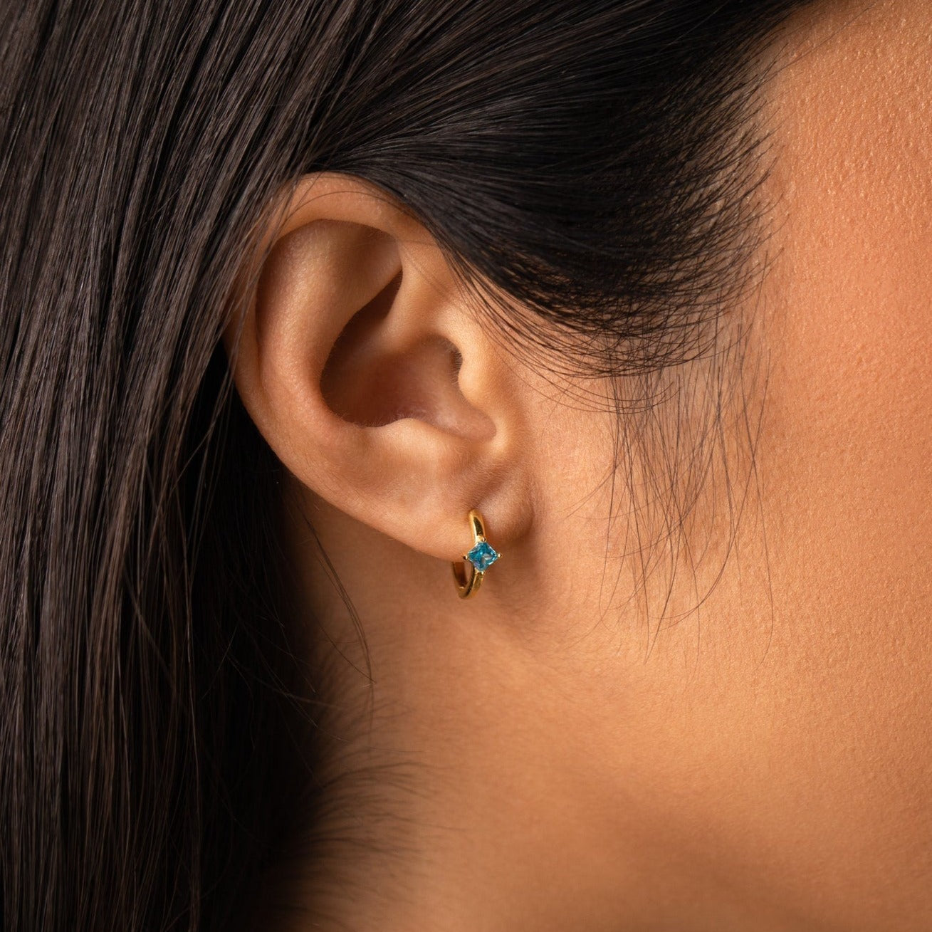 Close-up of a woman's ear wearing Athena Blue Zircon Huggies, gold hoop earrings with a striking blue gem—an ideal choice for fans of personalized jewelry.