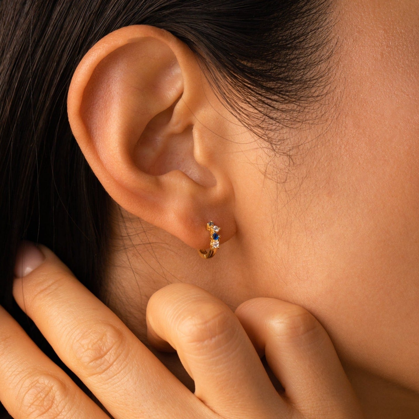 A close-up of an ear wearing the Angelica Sapphire Huggies, minimalistic gold hoops adorned with blue gemstones, as a hand gently touches the ear.