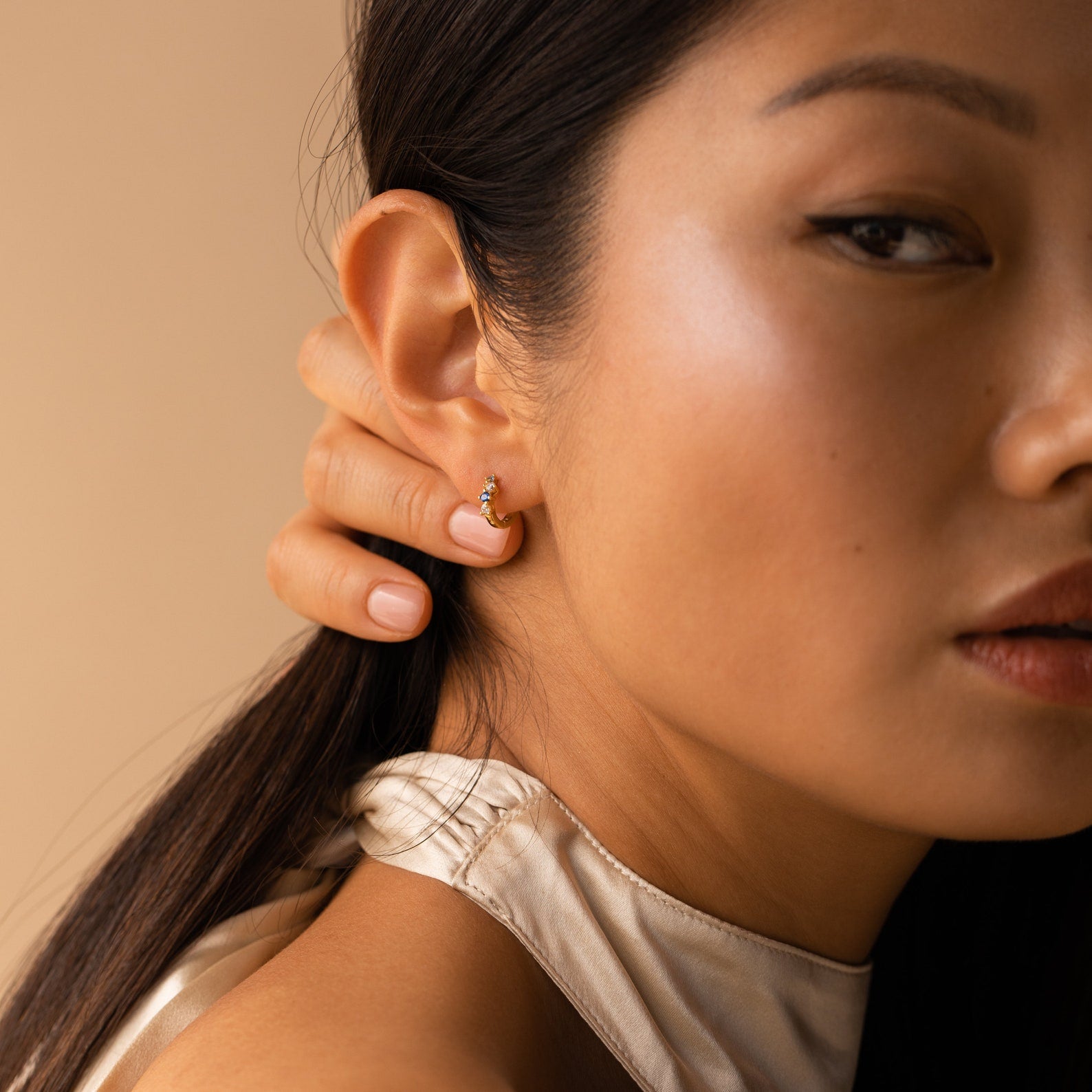 Woman with smooth skin and long dark hair models the Angelica Sapphire Huggies, elegant gold hoop earrings, paired with a satin top.