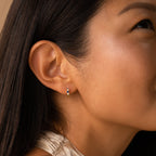 Close-up of a woman's ear wearing the Angelica Sapphire Huggies, minimalistic earrings with a blue gemstone stud; she has straight dark hair.