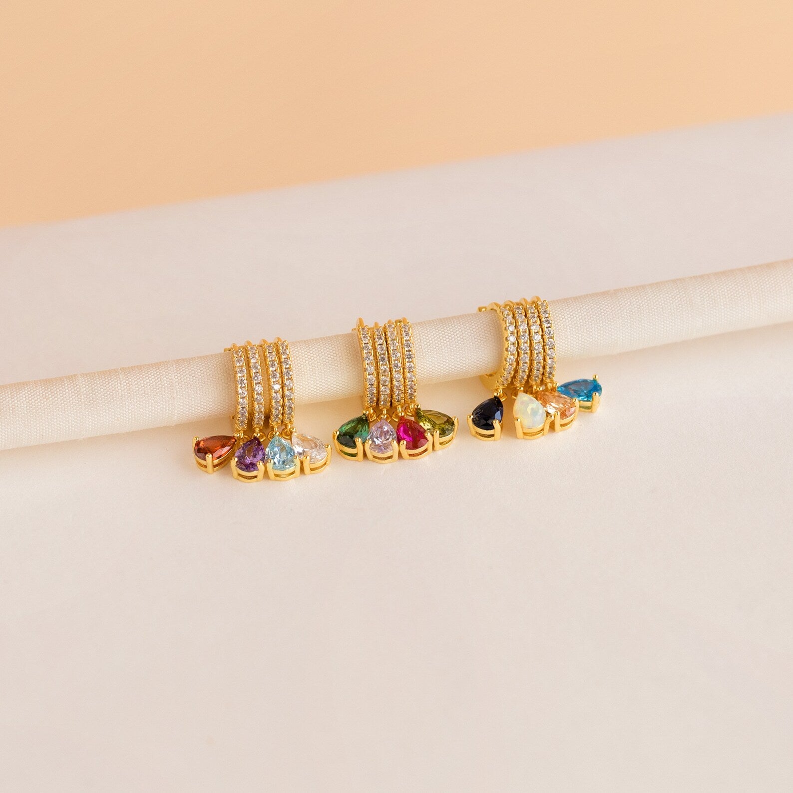 Four sets of Pave Birthstone Drop Huggies with colorful gemstones and pavé diamonds are elegantly displayed on a cream-colored rolled cloth.