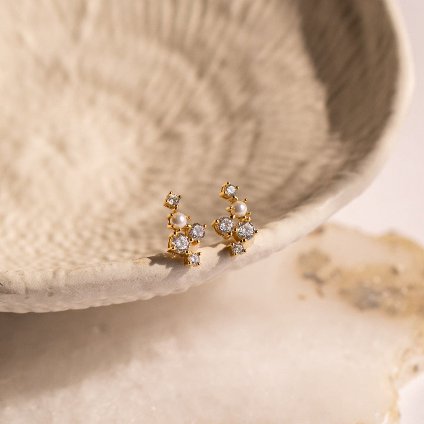 Gold and pearl stud earrings with sparkling diamonds, displayed on a textured ceramic dish—these Seine Diamond Studs are elegant wedding earrings featuring luminous cultured pearls.