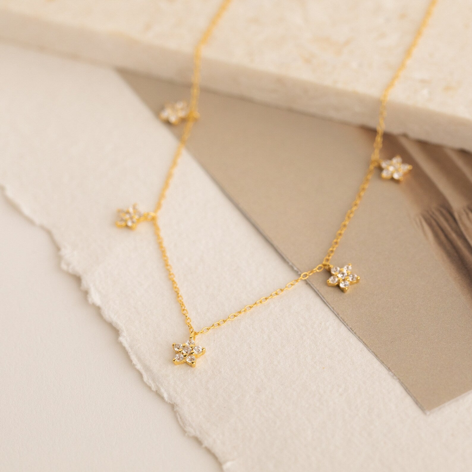 The Nanala Diamond Flower Necklace showcases gold, flower-shaped pendants, elegantly presented on textured neutral paper backgrounds for a refined, floral-inspired appearance.