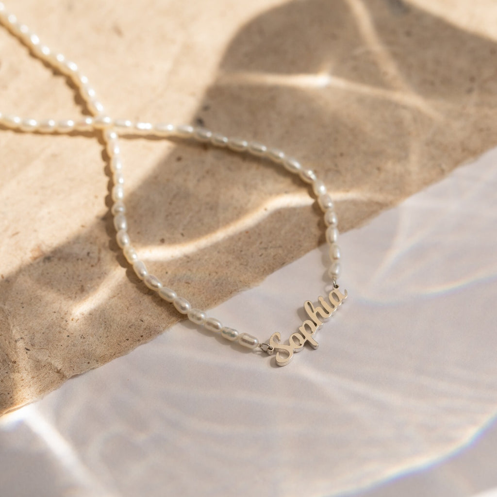 The Mellow Pearl Name Necklace features a delicate gold nameplate pendant accented with pearls, elegantly displayed on a textured, sunlit surface.