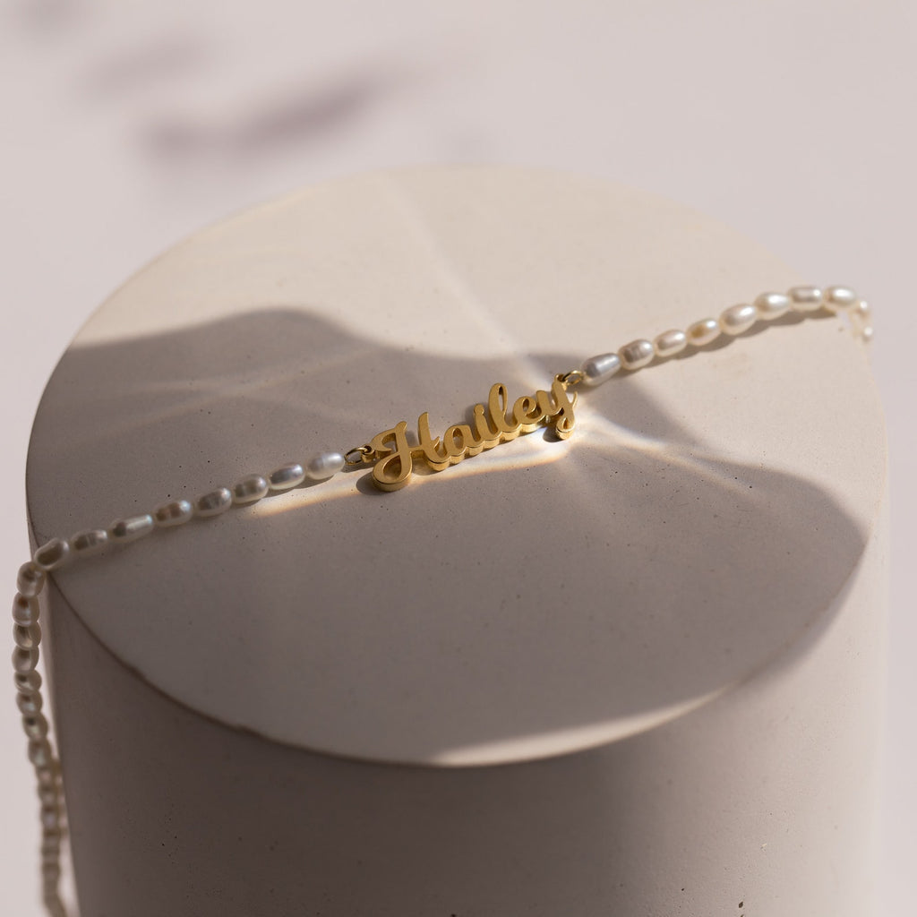 The Mellow Pearl Name Necklace features a gold dainty nameplate charm with pearl beads, elegantly displayed on a round, light-colored surface.