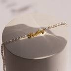 The Mellow Pearl Name Necklace features a gold dainty nameplate charm with pearl beads, elegantly displayed on a round, light-colored surface.