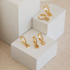 Pave Initial Huggies shown on white geometric blocks—stylish gold hoop earrings with customizable initial charms, making a perfect personalized gift.