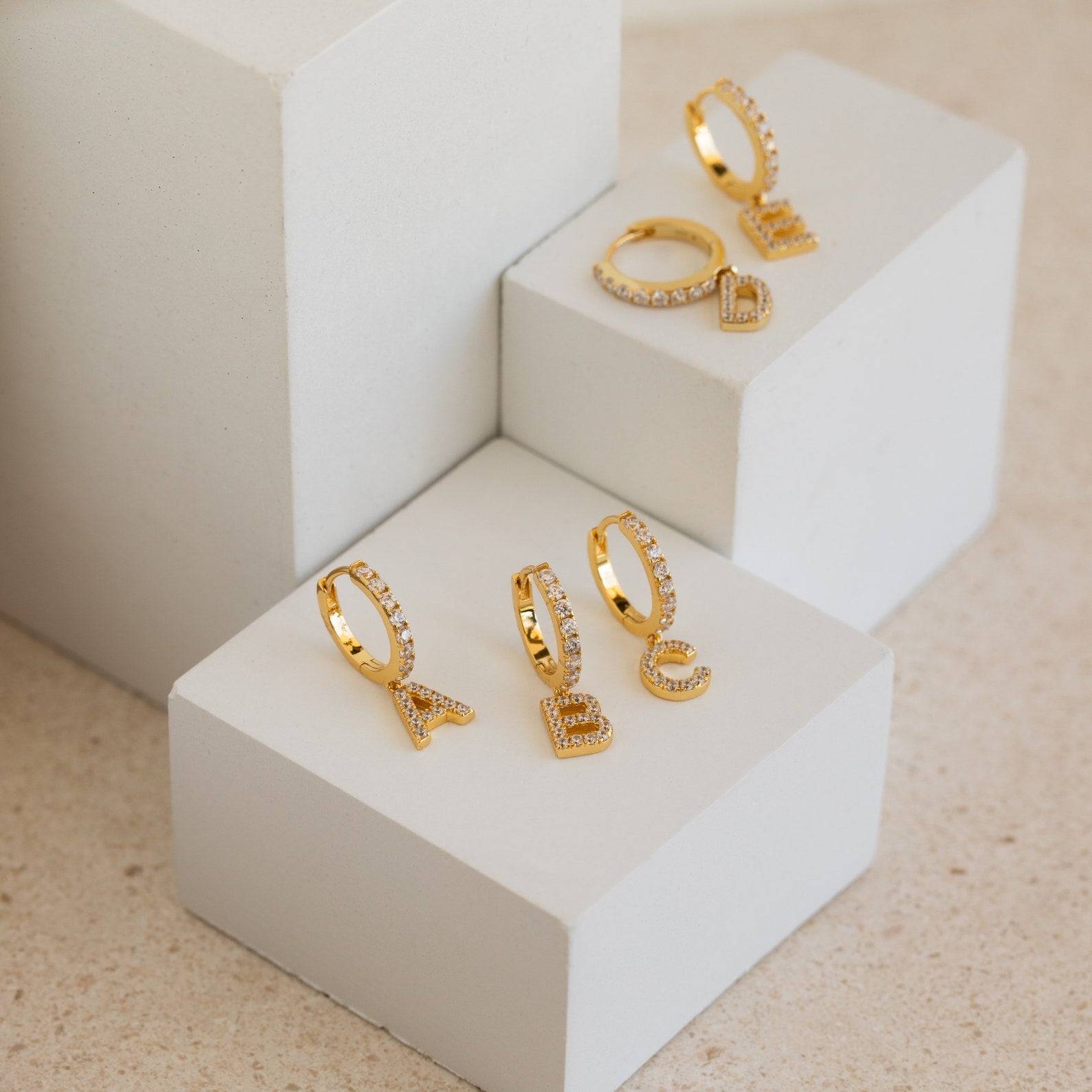 Pave Initial Huggies shown on white geometric blocks—stylish gold hoop earrings with customizable initial charms, making a perfect personalized gift.