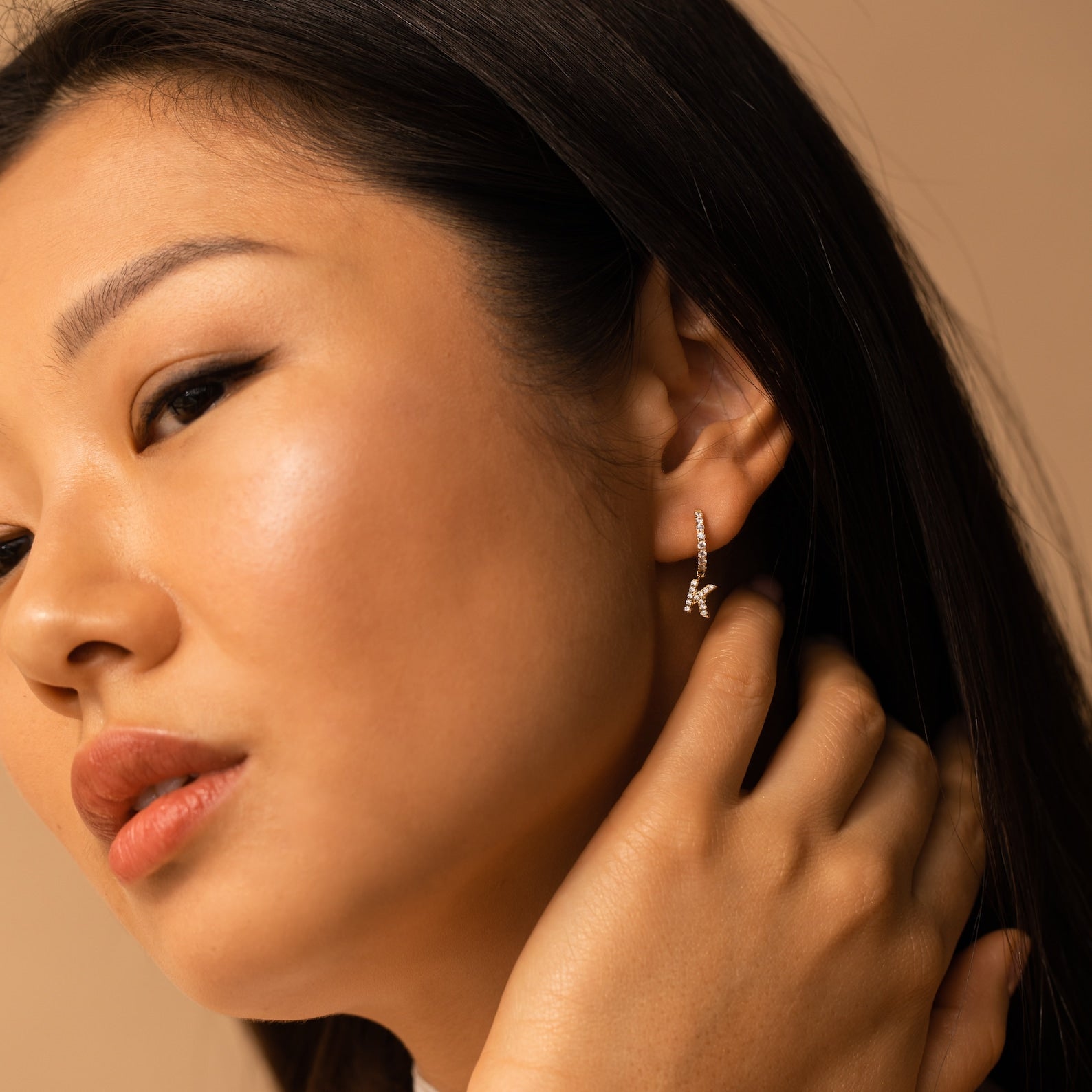 Close-up of a woman touching her ear, wearing the Pave Initial Huggies—small, elegant gold earrings shaped like the letter K—perfect as a personalized gift.
