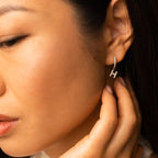 A woman touches her earring—a delicate gold hoop with sparkling stones. These Pave Initial Huggies make a stunning, personalized gift. Close-up on her ear and hand showcases the elegant detail.