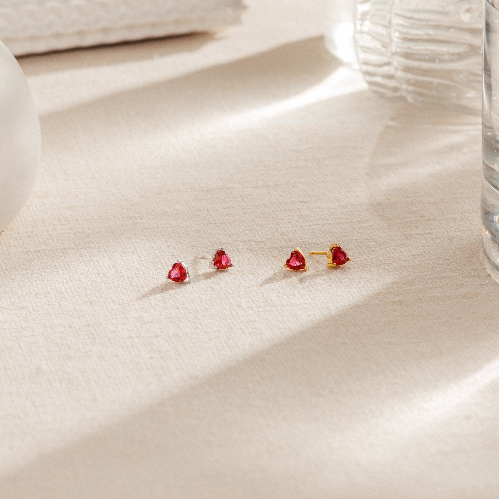 Ruby Heart Studs—gold earrings featuring small ruby heart-shaped studs displayed on a beige textured surface with soft lighting, perfect for adding a romantic touch to your date night look.