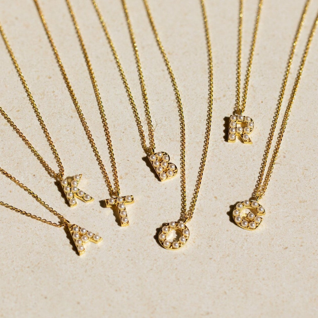 Seven gold necklaces with jeweled letter pendants—A, K, T, B, O, R, and I—are displayed on a beige background. Perfect for personalizing your look or layering with the Pearl Letter Necklace for a dainty pearls-inspired style.