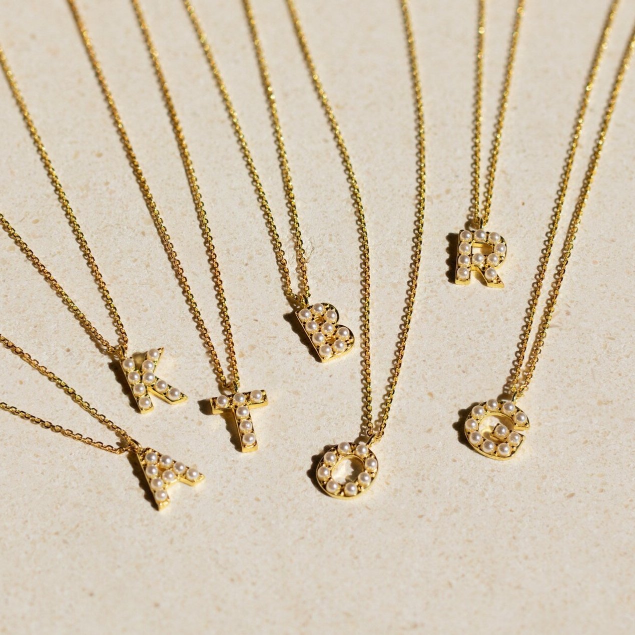 Seven gold necklaces with jeweled letter pendants—A, K, T, B, O, R, and I—are displayed on a beige background. Perfect for personalizing your look or layering with the Pearl Letter Necklace for a dainty pearls-inspired style.