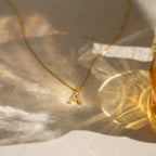 The Pearl Letter Necklace features a letter A pendant adorned with delicate pearls and gemstones, displayed on a sunlit surface—a beautiful piece of personalized jewelry.