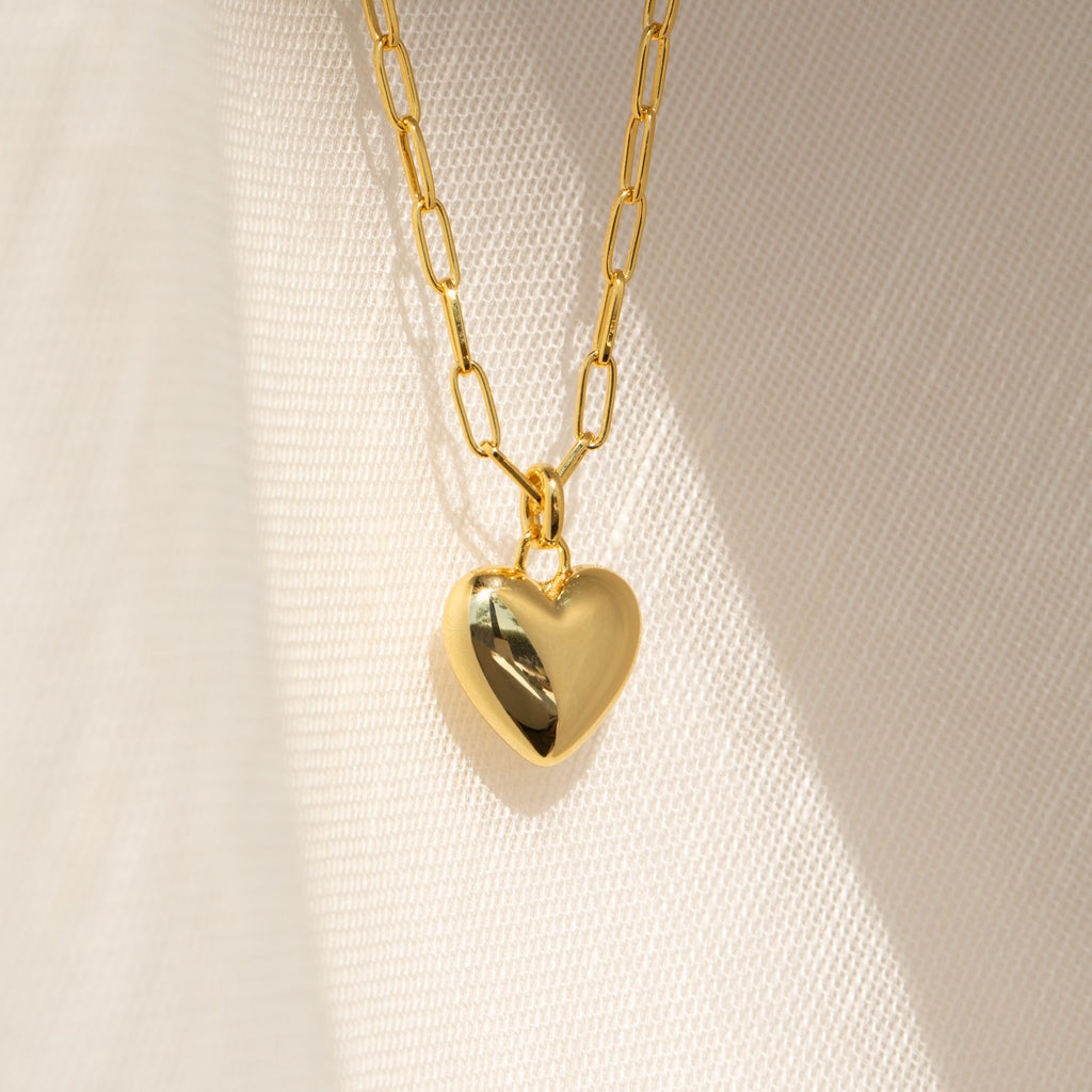 The Taylor Heart Paperclip Necklace features a gold heart locket pendant on a minimalist paperclip chain, displayed on a softly textured fabric background.