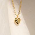 The Taylor Heart Paperclip Necklace features a gold heart locket pendant on a minimalist paperclip chain, displayed on a softly textured fabric background.