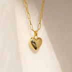 The Taylor Heart Paperclip Necklace features a gold heart locket pendant on a minimalist paperclip chain, displayed on a softly textured fabric background.