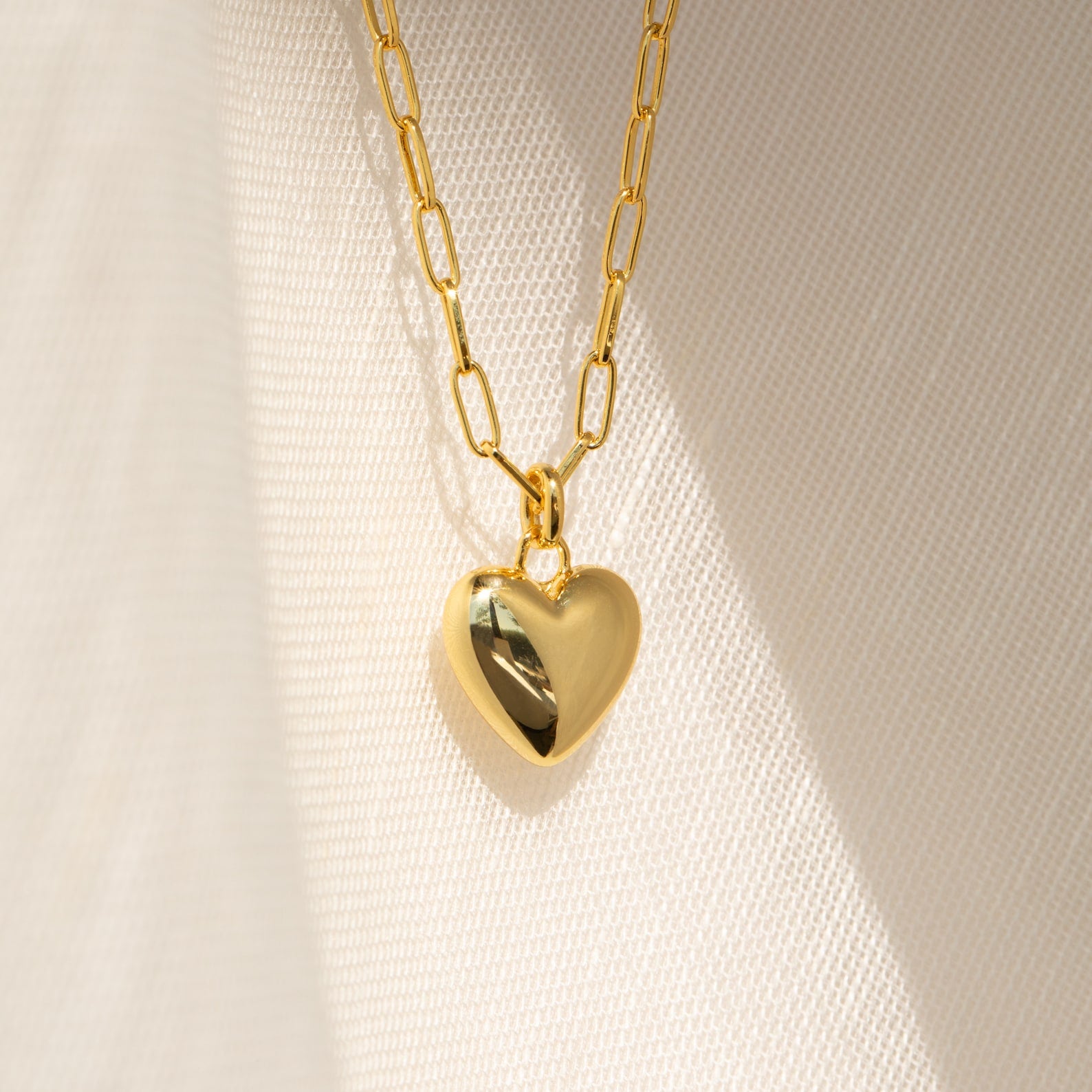 The Taylor Heart Paperclip Necklace features a gold heart locket pendant on a minimalist paperclip chain, displayed on a softly textured fabric background.