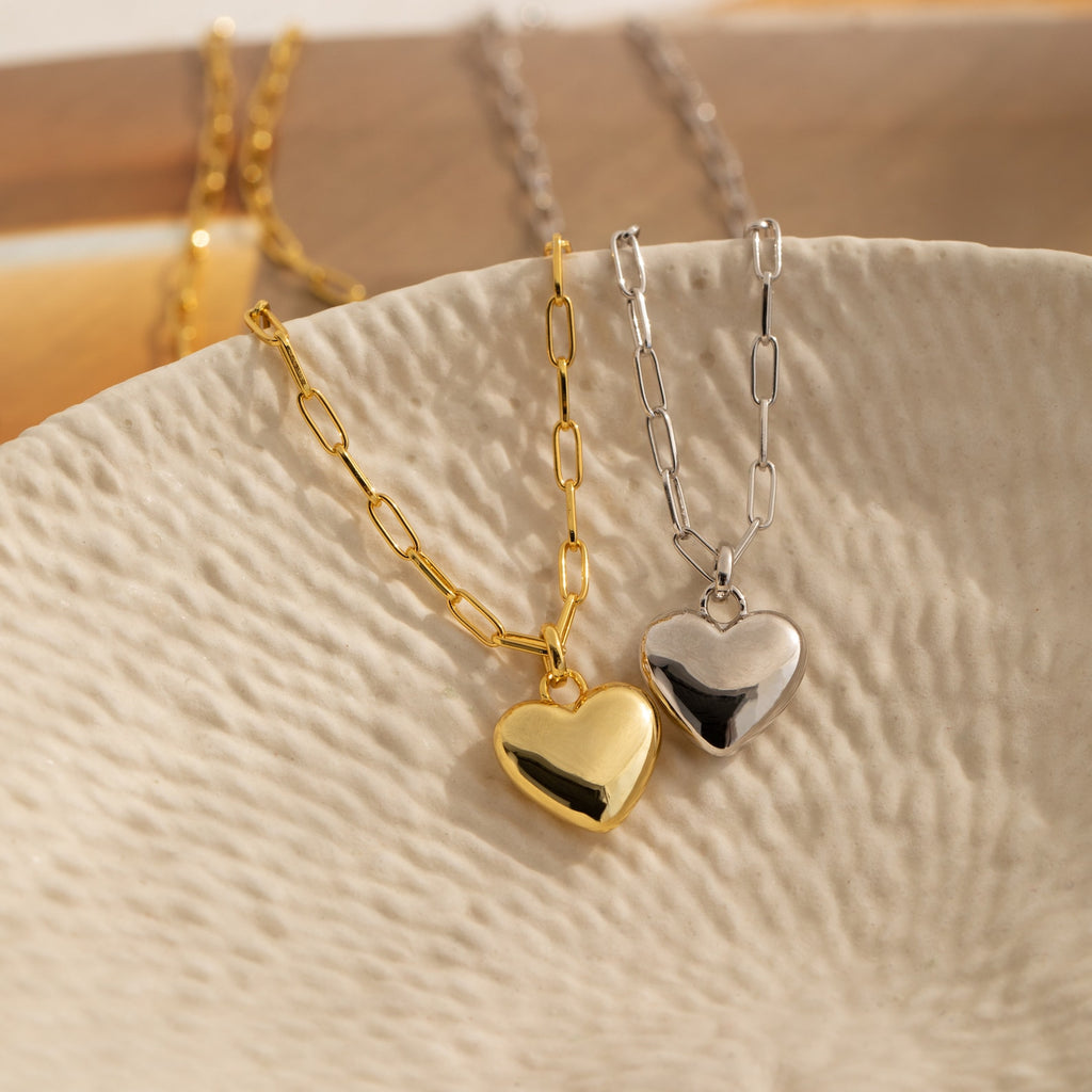 The Taylor Heart Paperclip Necklace, available in gold and silver, features a heart locket pendant and paperclip link chain, elegantly displayed on a textured cream dish.