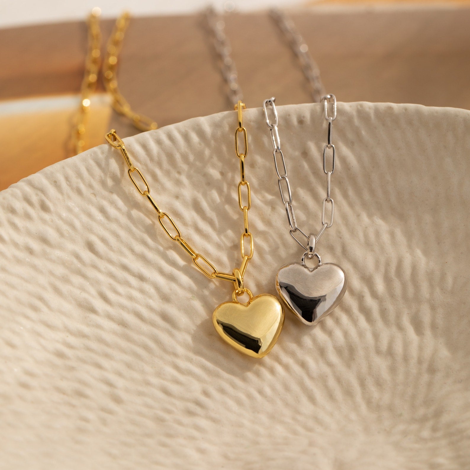 The Taylor Heart Paperclip Necklace, available in gold and silver, features a heart locket pendant and paperclip link chain, elegantly displayed on a textured cream dish.
