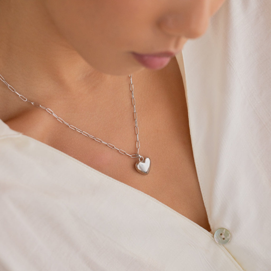 A woman shown from the shoulders down wears a white blouse and the Taylor Heart Paperclip Necklace, featuring a silver heart-shaped pendant on a paperclip link chain.