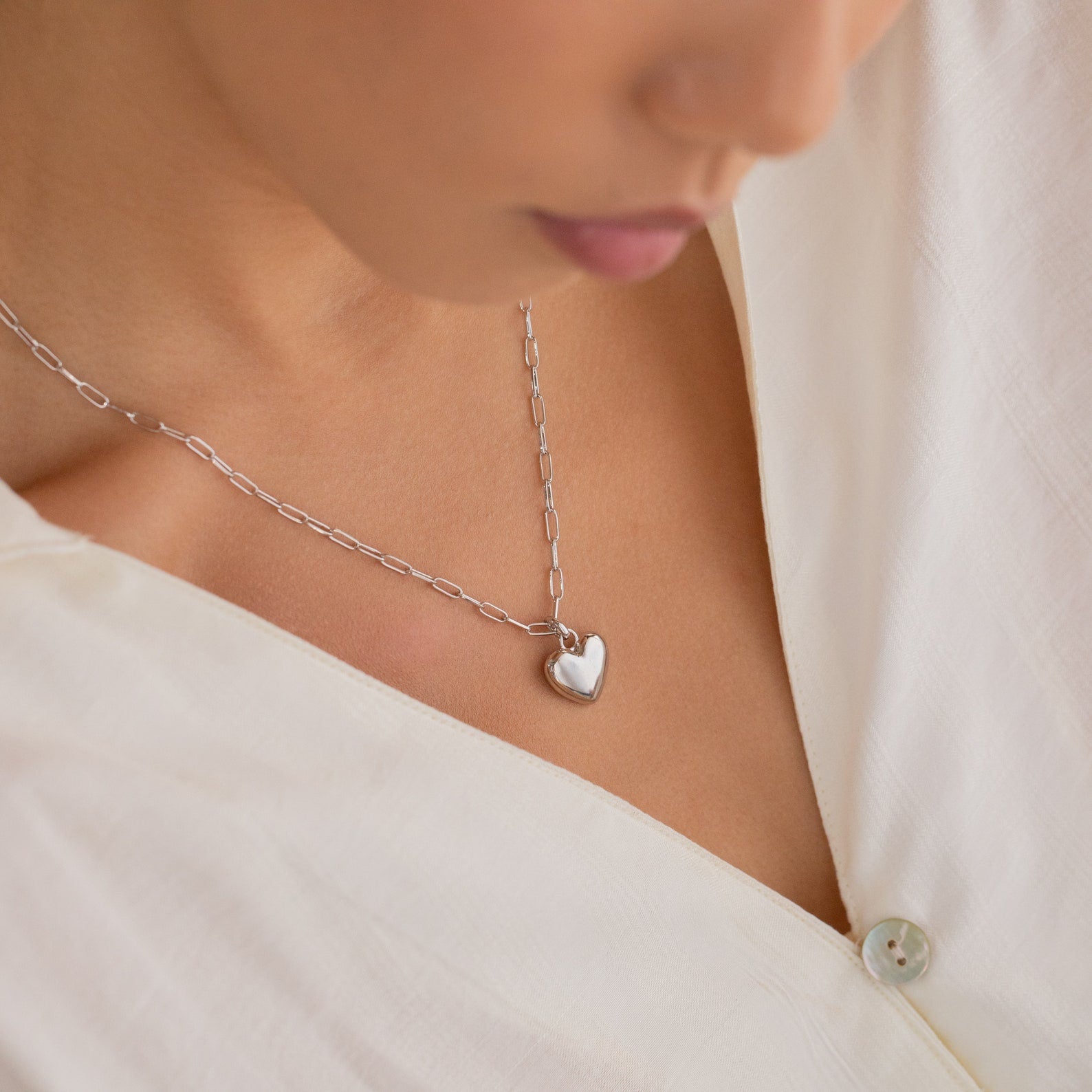 A woman shown from the shoulders down wears a white blouse and the Taylor Heart Paperclip Necklace, featuring a silver heart-shaped pendant on a paperclip link chain.