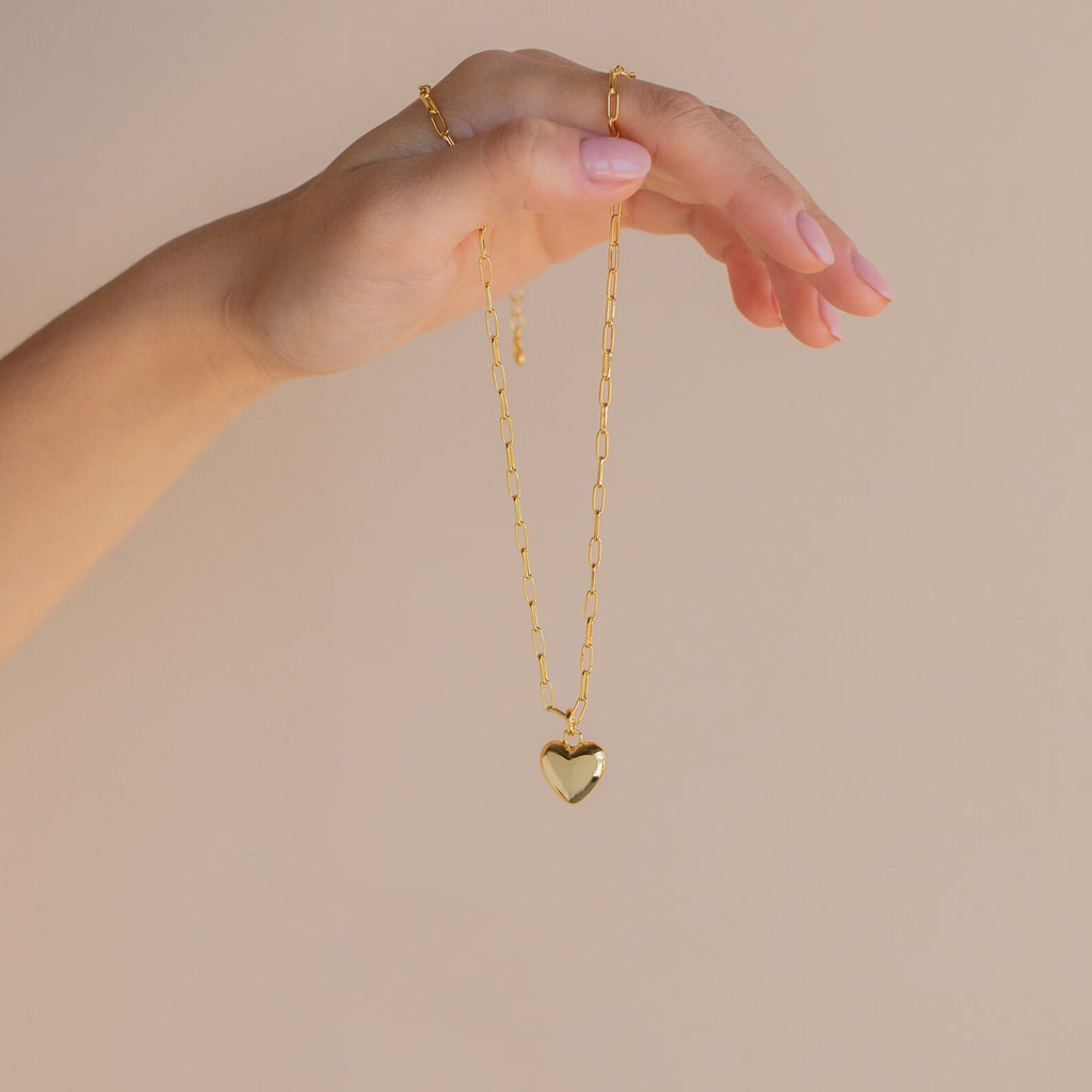 A hand delicately holds the Taylor Heart Paperclip Necklace, featuring a shiny heart-shaped pendant on a gold chain, against a beige background.