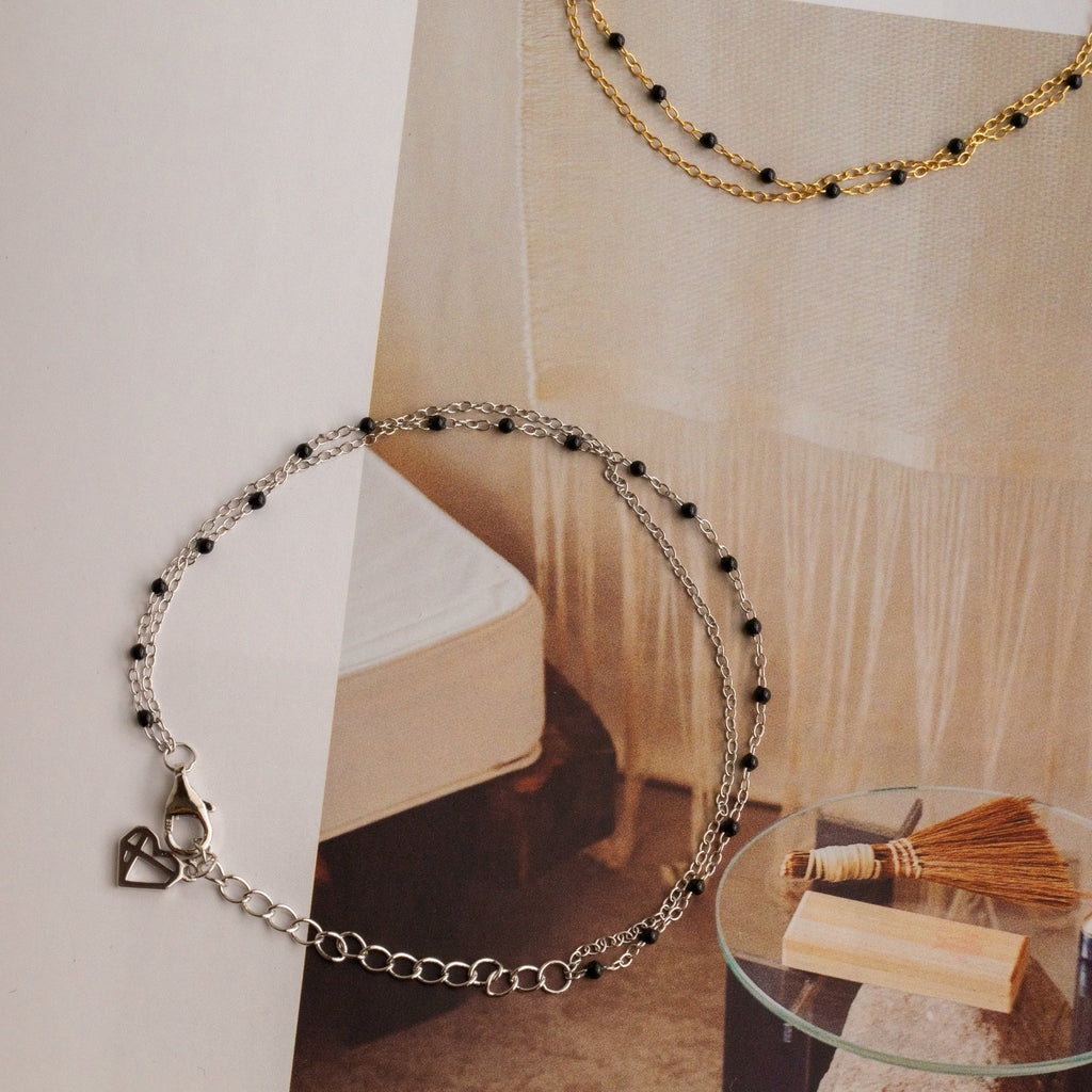 The Calypso Station Bracelet features a delicate silver chain with small black beads, ideal for stacking or pairing with boho accessories. Shown on a decorative background alongside a gold chain.