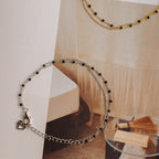 The Calypso Station Bracelet features a delicate silver chain with small black beads, ideal for stacking or pairing with boho accessories. Shown on a decorative background alongside a gold chain.