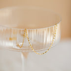 Delicate gold chain necklace with small faceted links draped over the rim of a clear ribbed glass coupe.
