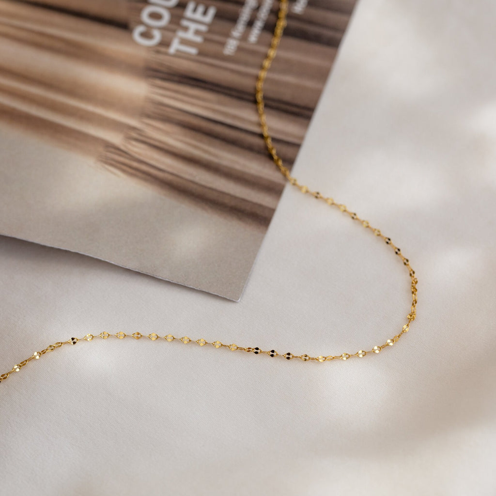 The Josephine Chain Necklace, a delicate gold mirror chain perfect for wedding jewelry, is displayed on a white surface next to a magazine.