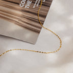 The Josephine Chain Necklace, a delicate gold mirror chain perfect for wedding jewelry, is displayed on a white surface next to a magazine.
