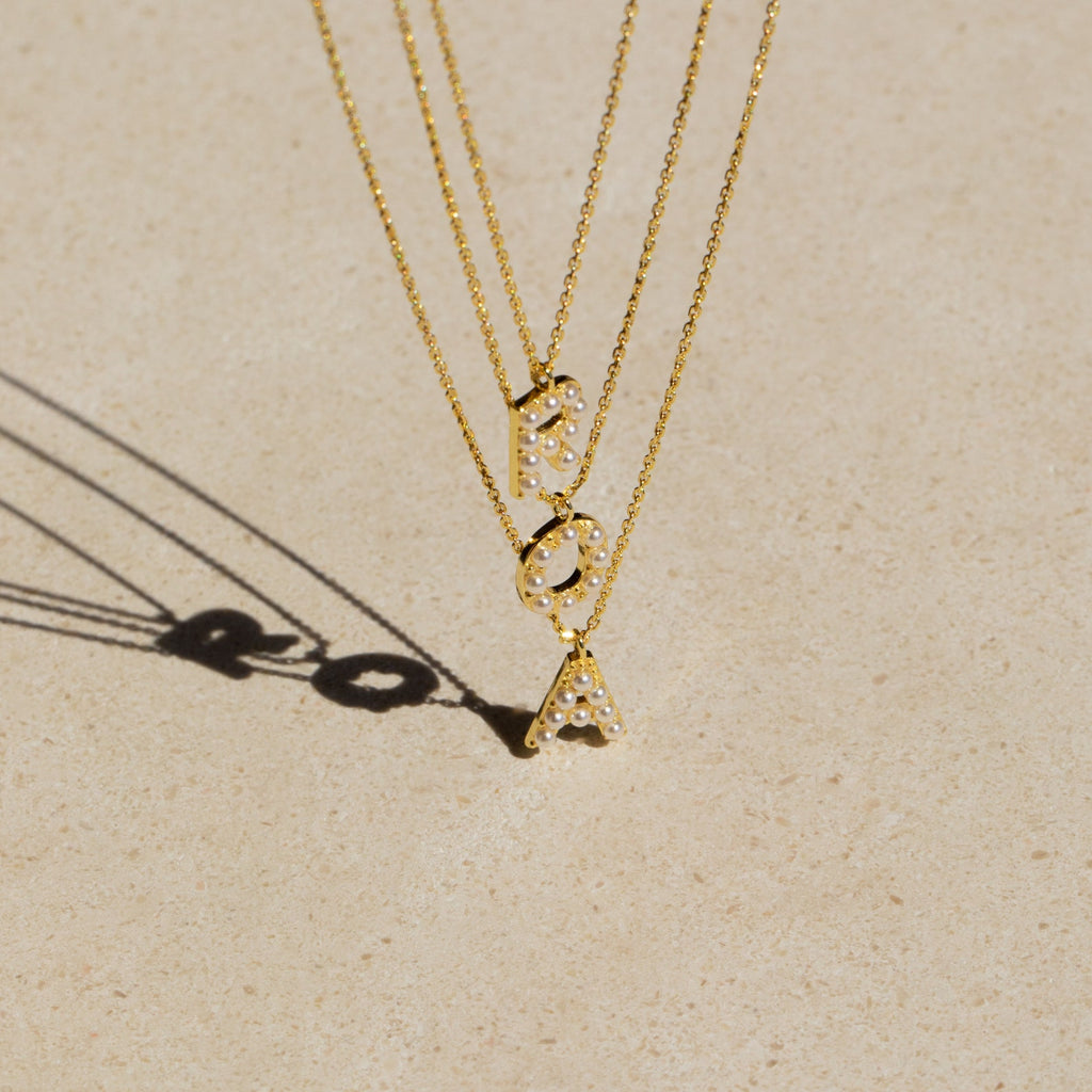 The Pearl Letter Necklace features gold pendants with the letters R, O, and A accented by dainty pearls on a beige surface—ideal for fans of personalized jewelry.