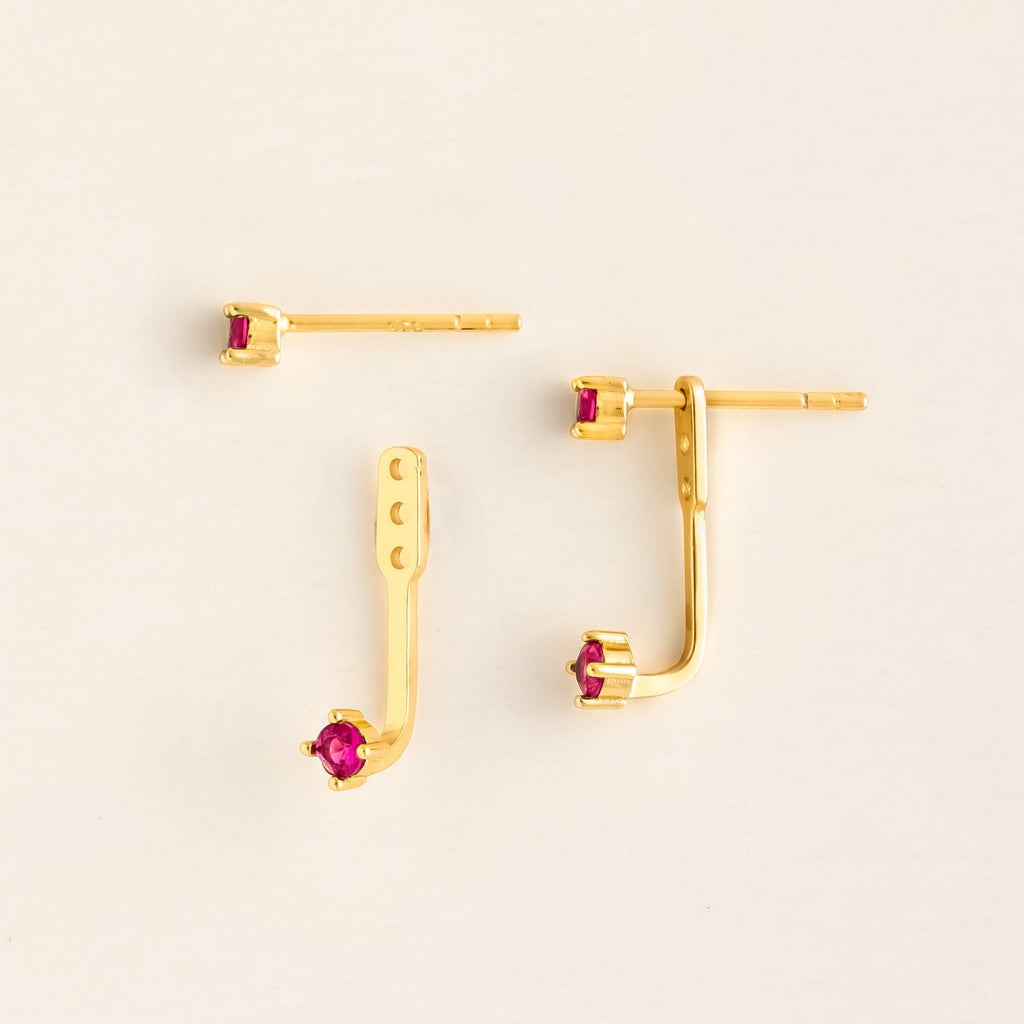 Flat lay of a pair of gold ear jacket earrings with bright Ruby round-cut gemstones. The set includes a small front stud and a curved backing piece with adjustable holes, designed to peek from behind the earlobe.