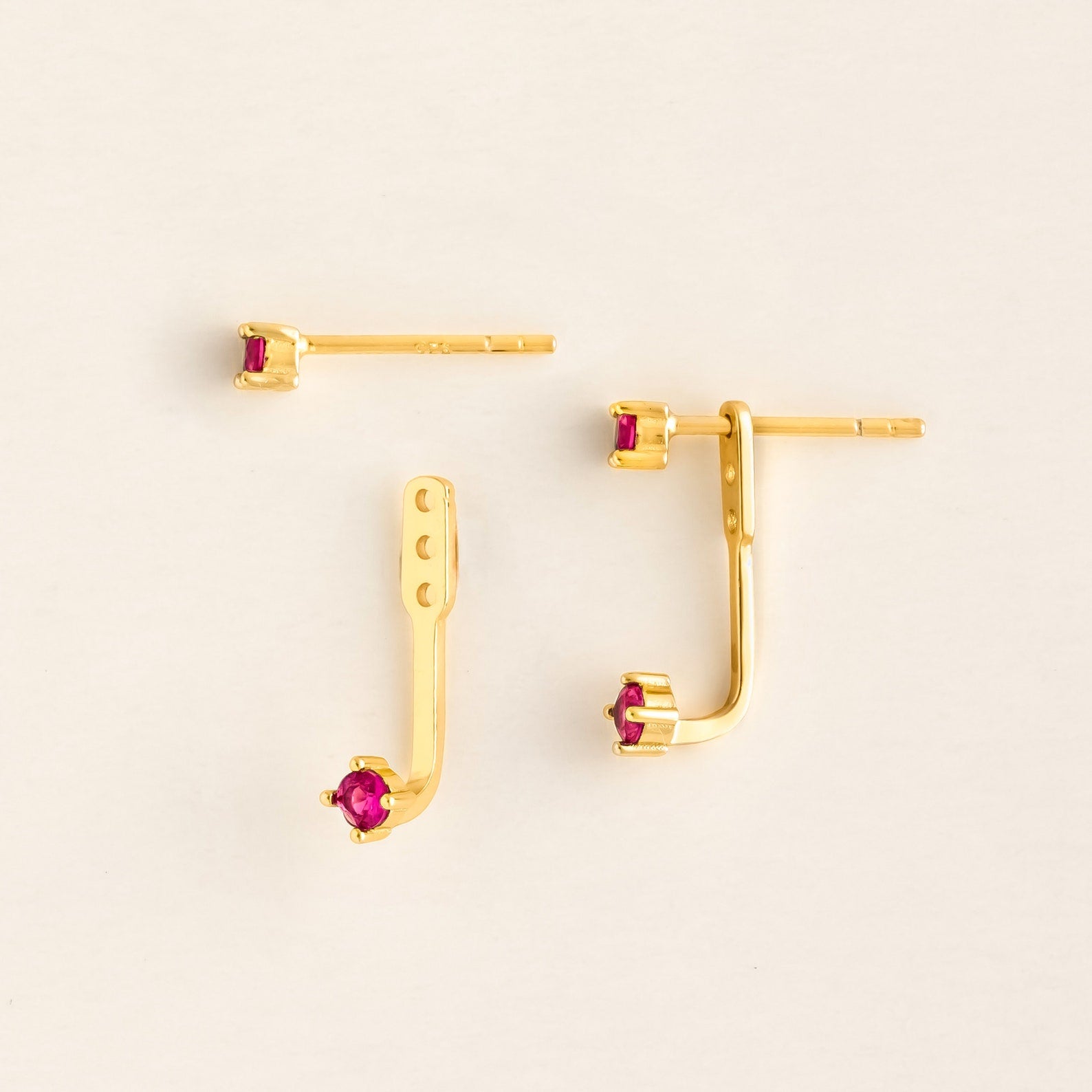Flat lay of a pair of gold ear jacket earrings with bright Ruby round-cut gemstones. The set includes a small front stud and a curved backing piece with adjustable holes, designed to peek from behind the earlobe.
