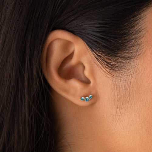 Close-up of a woman's ear wearing Bella Birthstone Studs—gold earrings with three small blue gemstones, ideal personalized jewelry for mothers and daughters.