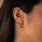 Close-up of a woman’s ear wearing the Pave Birthstone Drop Huggies—pavé diamond hoop earrings featuring a green teardrop-shaped gemstone charm.