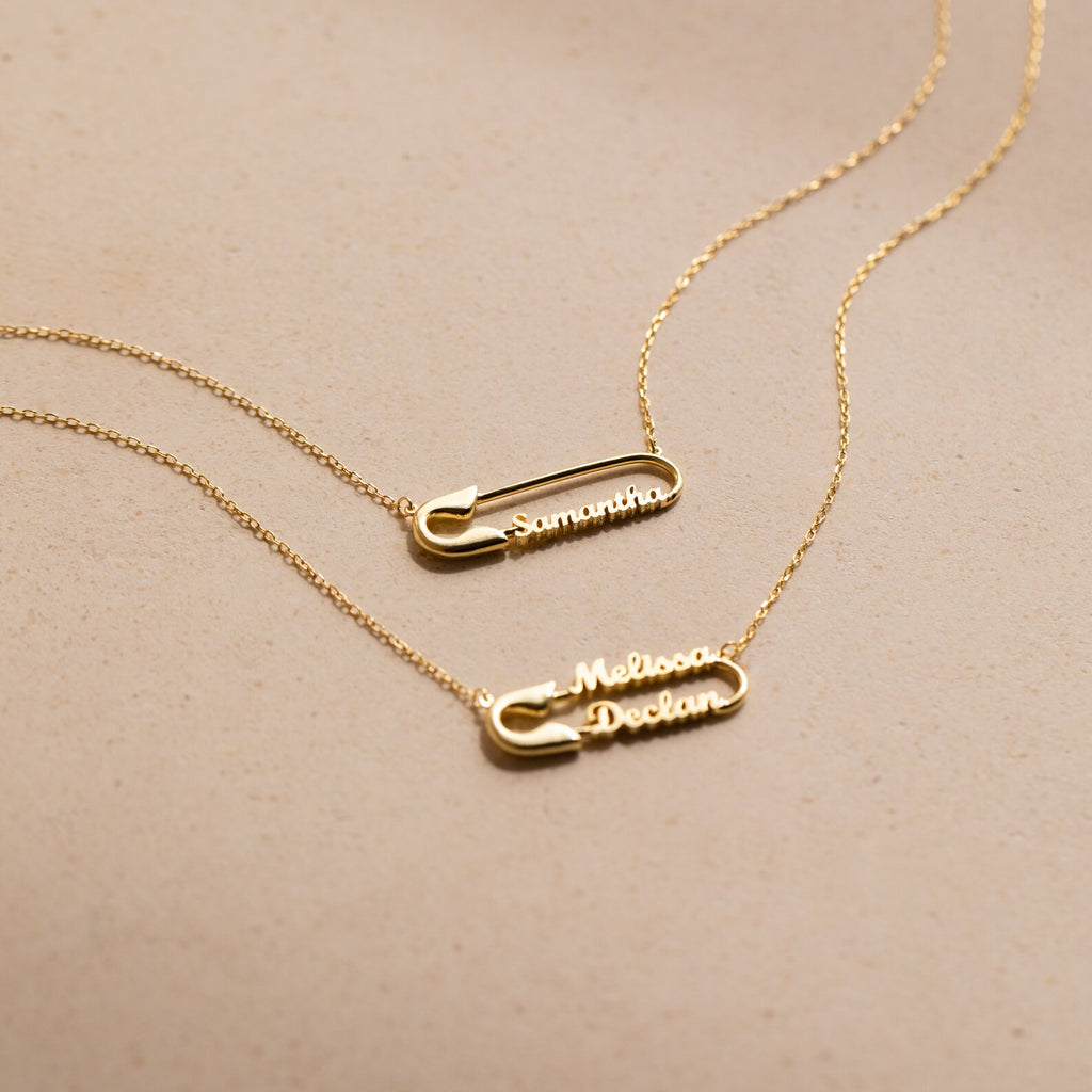 Two gold Safety Pin Name Necklaces personalized with the names Samantha and Melissa Declan make a chic statement accessory against a beige background.