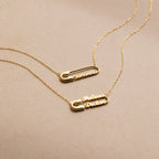Two gold Safety Pin Name Necklaces personalized with the names Samantha and Melissa Declan make a chic statement accessory against a beige background.