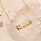 The Safety Pin Name Necklace features a striking gold pendant and rests elegantly on a light stone surface, making it the perfect statement accessory.