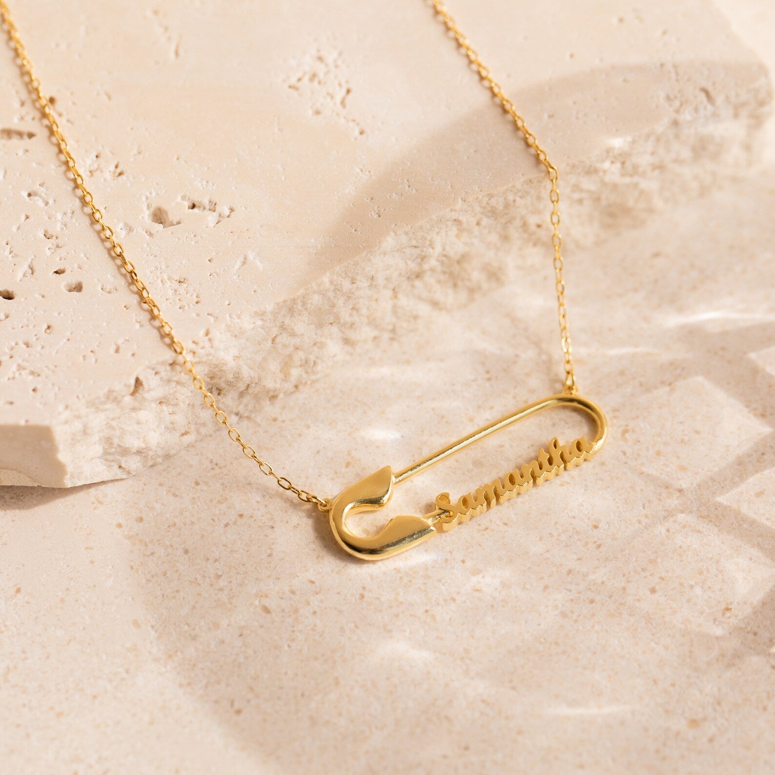 The Safety Pin Name Necklace features a striking gold pendant and rests elegantly on a light stone surface, making it the perfect statement accessory.