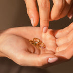 A hand displays three gold rings with vibrant gemstones as another selects the Athena Opal Huggies.