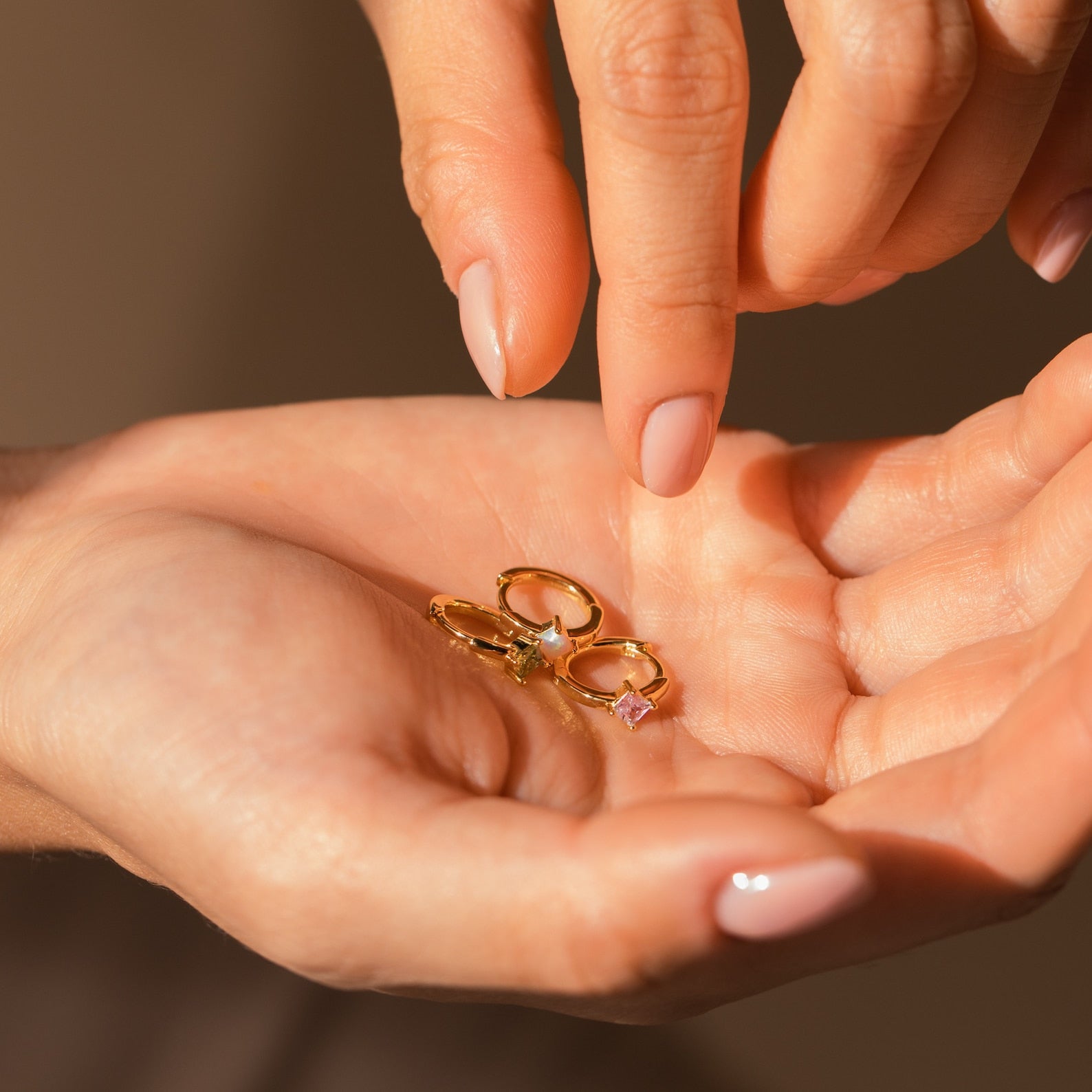 A hand displays three gold rings with vibrant gemstones as another selects the Athena Opal Huggies.