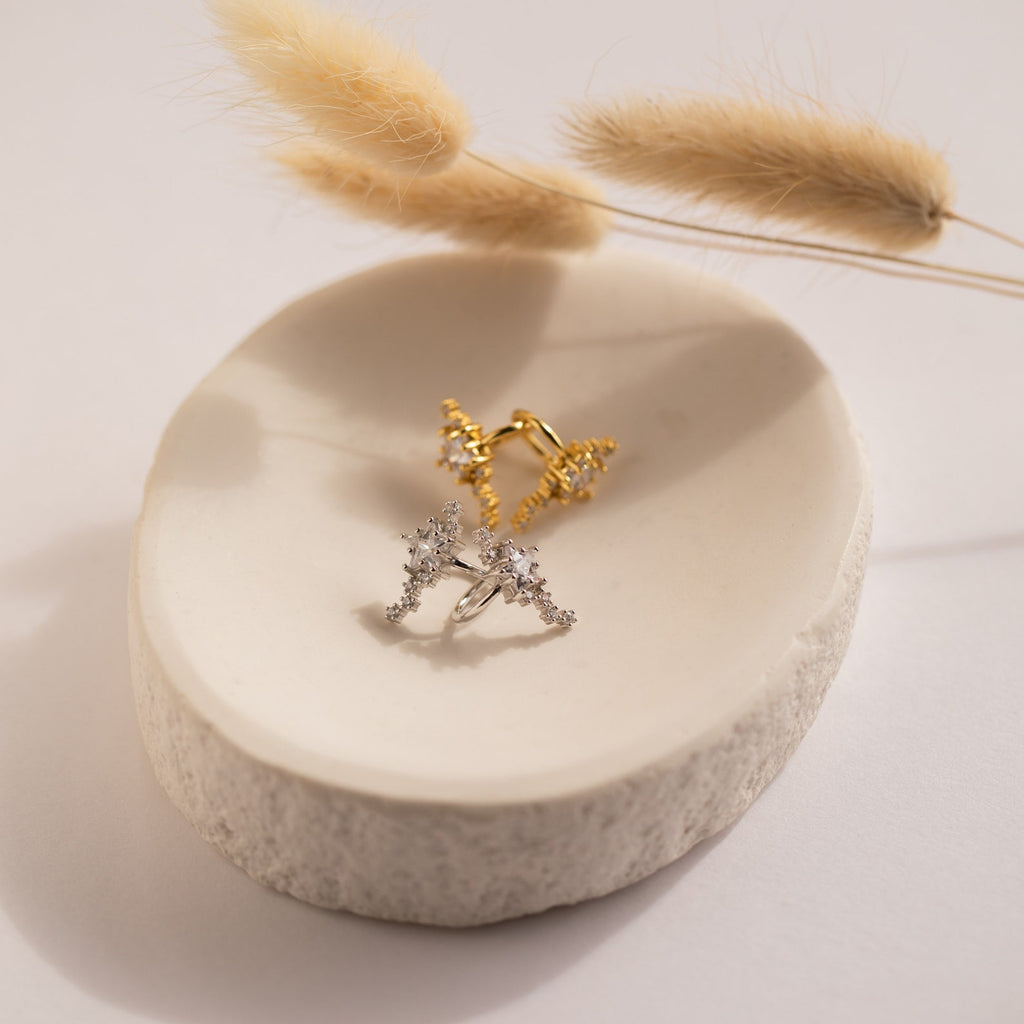 Gold and silver rings with crystal accents rest on a white ceramic dish beside dried grasses. The Starry Night Ear Cuffs, designed for a no-piercing look, complete this elegant display.